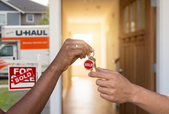 Two hands—one of a Black woman with a wedding ring and one multi-ethnic—handing over a set of house keys with a bright red 'SOLD' tag. The background shows a clear 'SOLD' sign in the yard, a moving truck, and the open door of a modern house.